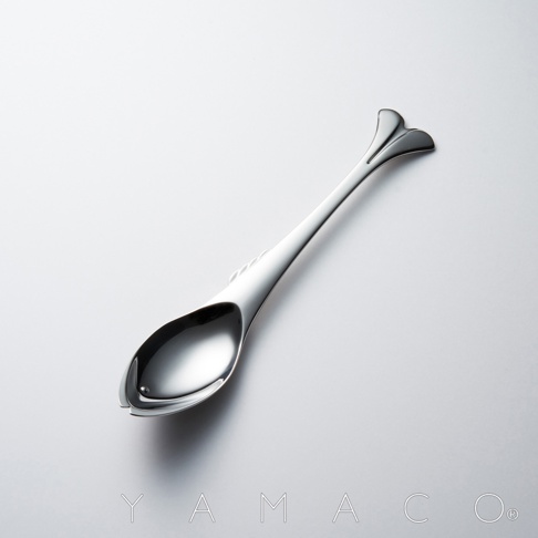 YAMACO 品牌標誌，一隻銀色不鏽鋼湯匙，湯匙頭設計成魚的形狀，湯匙柄末端有魚鰭造型，背景為純白色。