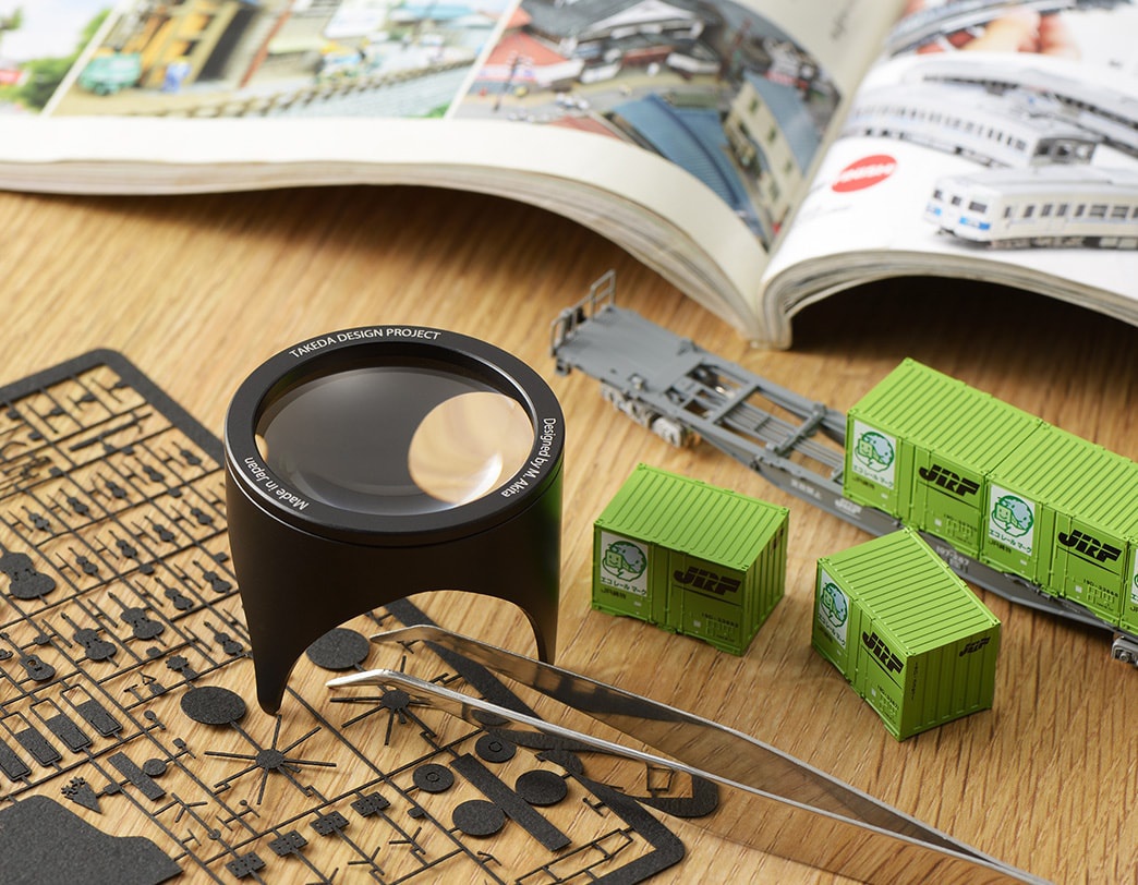 黑色鋁製閱讀放大鏡，鏡筒上有「TAKEDA DESIGN PROJECT」及「Made in Japan」字樣，下方有兩支鑷子，旁邊散落著黑色模型零件板，背景是打開的書籍和綠色貨櫃模型。