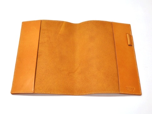 職人手作皮革A5筆記本書套，呈現自然皮革紋理，顏色為溫暖的橘棕色，內裡為絨面處理。書套兩側有皮革夾層，右側邊緣有細緻的縫線，右下角印有「HERZ」品牌標誌。