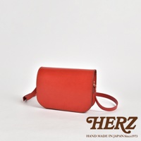 一個紅色的皮革翻蓋橫式單肩包，配有可調節的肩帶，包身線條簡潔，縫線清晰，呈現職人手作質感。