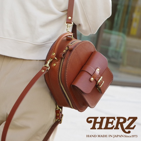 咖啡色皮革迷你後背包，圓形主體搭配前方方形口袋，設有拉鍊開合及金屬扣環，可肩背或手提。