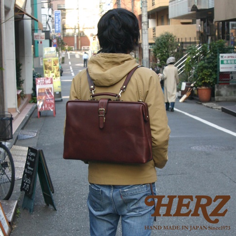 職人手作的咖啡色皮革軟式杜勒斯包，帶有金色金屬釦環和提把，肩背帶連接至背包。