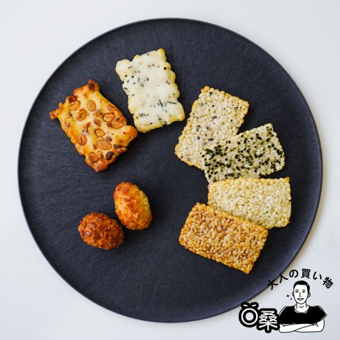 黑盤上擺放著多種米菓，包含花生米菓、點綴海苔的米菓、以及原味和焦糖色米菓，還有兩顆金黃色的炸米球。