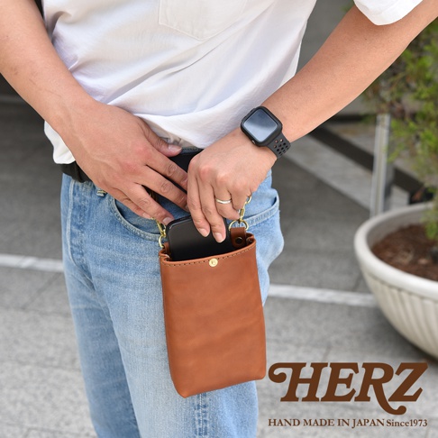 職人手作皮革兩用手機包，展示一款棕色皮革手機套，可掛於腰間，搭配牛仔褲與白色上衣。