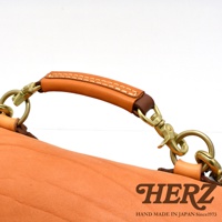 職人手作皮革鹿角扣繩郵差包，特寫皮革提把與黃銅五金扣環，包身為淺棕色皮革，提把處縫線明顯。