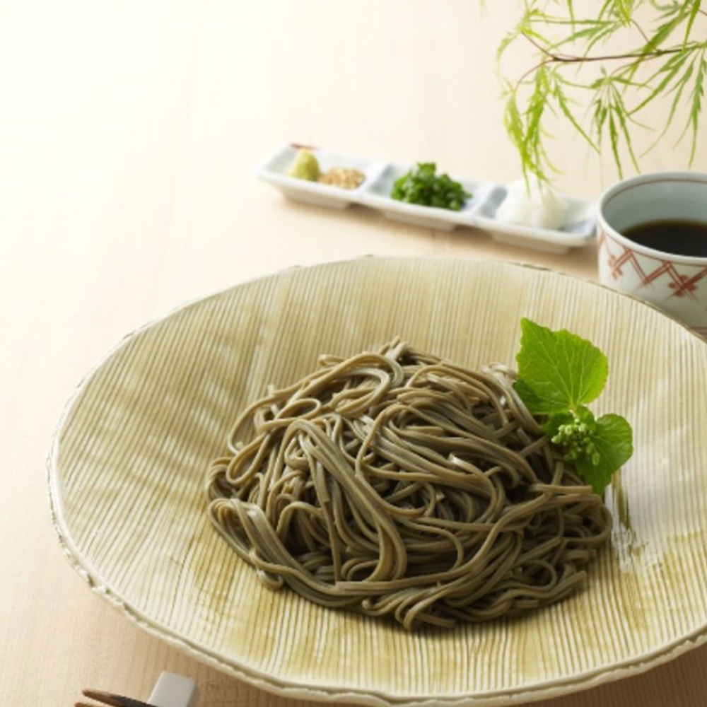 一盤盛著濃郁綠色海苔蕎麥麵，麵條細緻捲曲，擺放在帶有紋路的淺綠色盤子中央，旁邊點綴著一抹鮮綠的葉子。背景處有裝有醬汁的小杯和三格小碟，分別盛有芥末、蔥花和薑末。