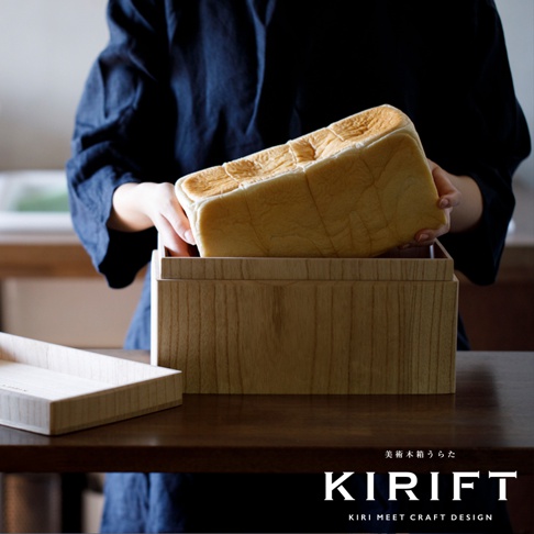 淺色桐木製長方形麵包保存箱，箱蓋上方放置著一條金黃色表皮的方形吐司麵包，雙手正將吐司放入箱內。