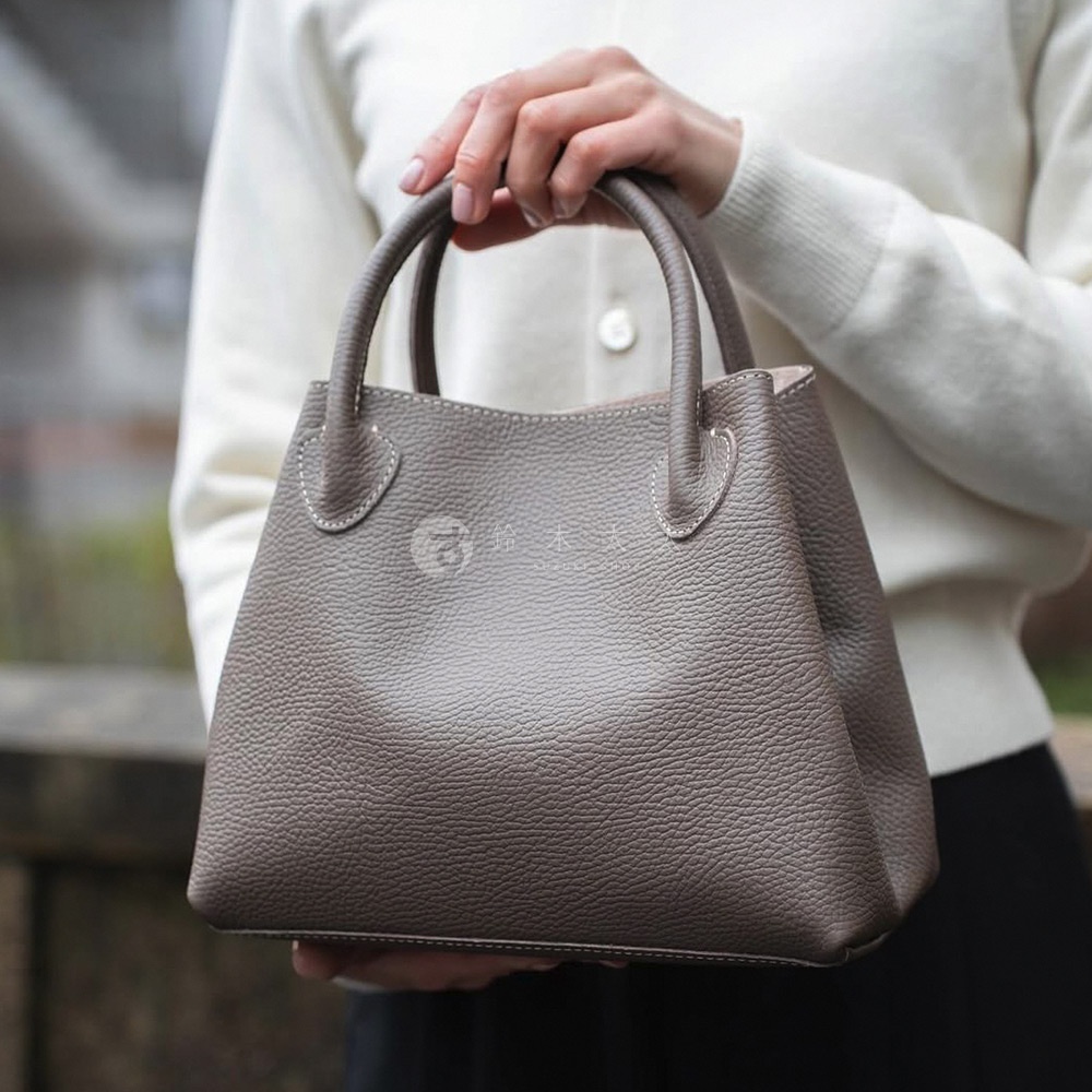 一位女士手持一個灰褐色、荔枝紋皮革材質的兩用手提托特包，包包結構挺括，配有圓潤的手提把，縫線清晰可見。