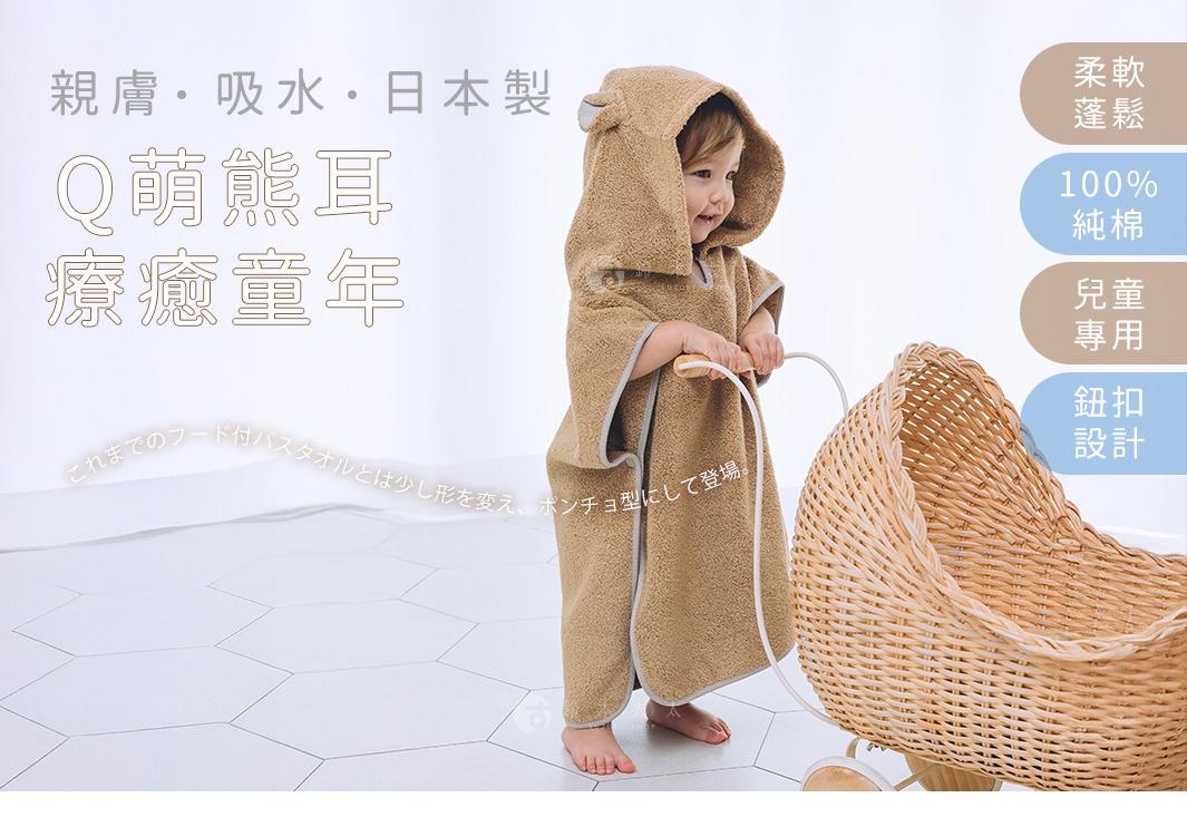 一位可愛的幼童穿著一件棕色、帶有熊耳朵的斗篷式連帽浴巾，浴巾為純棉材質，觸感柔軟蓬鬆，適合兒童使用，並有鈕扣設計。