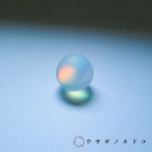 霧面質感的透明玻璃球，內部折射出橙色和粉色的光暈，置於淺藍色背景上，球下方投射出綠色的陰影。