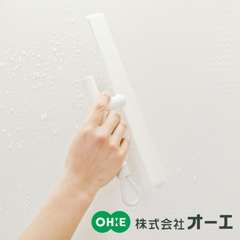 一只手正握著白色刮水刷，在帶有水珠的白色紋理牆面上使用。刮水刷由白色塑膠製成，握把部分為圓柱形，刮片為扁平長條狀。