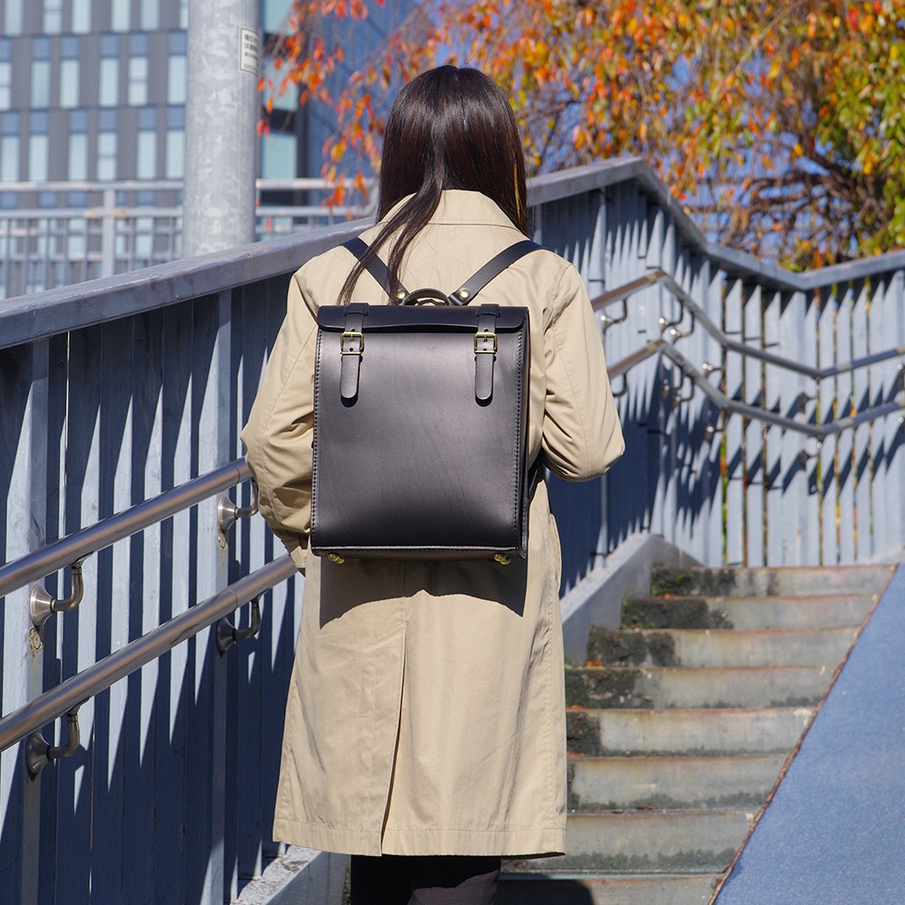 一名女子背著一個黑色皮革後背包，包款為方形設計，有兩個皮帶扣和金屬扣環。