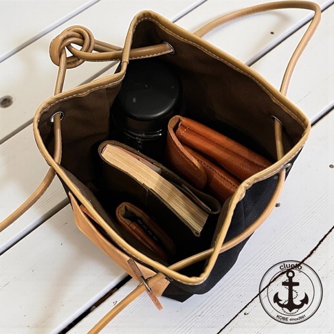 神戶clueto品牌義大利皮革束口肩背包，內部裝有錢包、筆記本和黑色瓶子，背景為白色木板