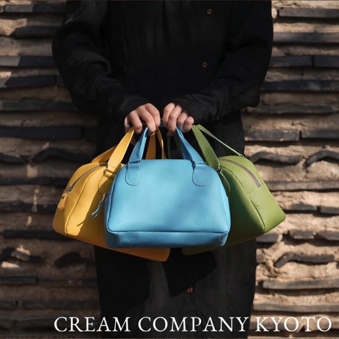 三人各持一個半月形狀的皮革迷你手提包，顏色分別為亮黃色、天藍色和橄欖綠。包身呈現細緻的荔枝紋皮革質感，設計簡約，配有短提把。