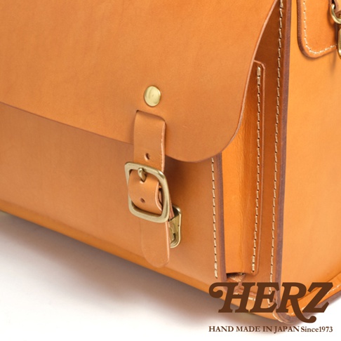 HERZ 品牌標誌，下方為 HAND MADE IN JAPAN Since1973，圖片為淺棕色皮革橫式書包的特寫，展示了金屬釦環和縫線細節