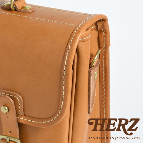 職人手作皮革商務休閒兩用單肩包，採用淺棕色皮革，搭配米白色縫線，包款有翻蓋設計，配有金屬扣環和提把，整體展現經典質感。