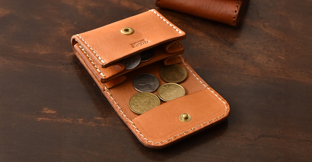 這款日本HERZ職人手作皮革零錢包呈現淺棕色，採雙折式設計並配有金屬按扣，包內印有HERZ標誌與MADE IN JAPAN字樣，質感厚實，內部空間可收納多枚硬幣。