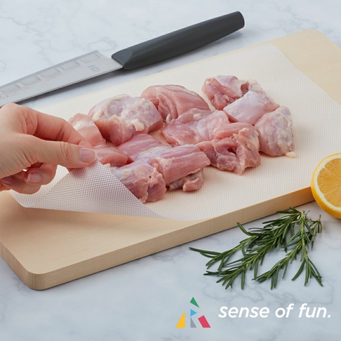 捲筒式砧板保潔墊片，半透明材質，帶有網格紋理，鋪設在木質砧板上，上方擺放著切塊的生雞肉、一把刀、檸檬和迷迭香。