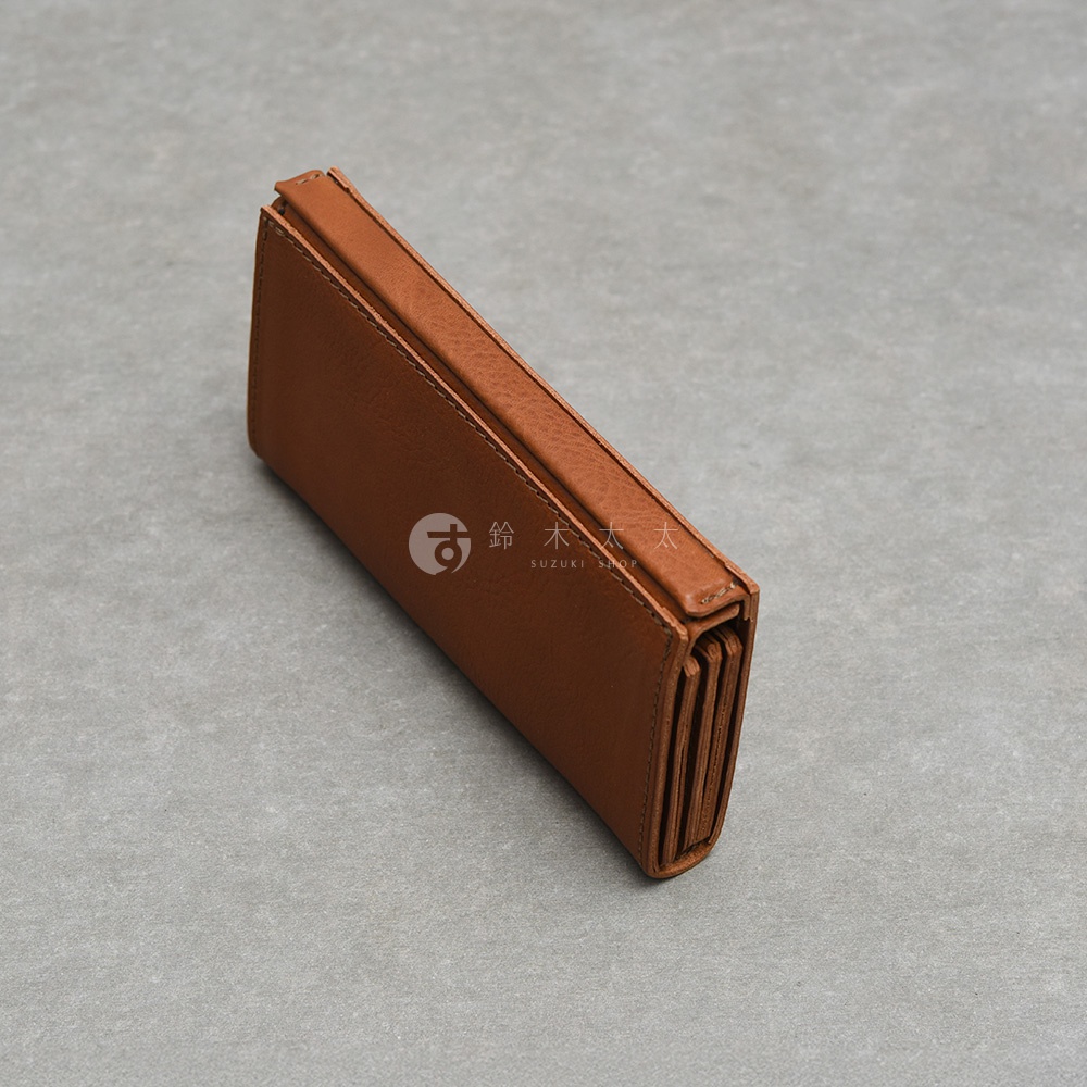 這是一個日本HERZ職人手作皮革長夾，採用暖棕色植鞣革製成，呈現簡約洗練的細長外觀。側面可見獨特的風琴摺疊結構，展現出色的收納空間與工藝細節。夾身表面帶有鈴木太太SUZUKI SHOP的淡色浮水印標誌，整體造型沉穩且具備優質皮革的自然紋理質感。