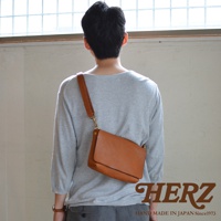 一個穿著灰色上衣的人斜背著棕色皮革包，包包的款式簡約，皮革材質看起來質感良好。