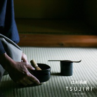 一手捧著黑色抹茶碗，碗中置有竹製抹茶茶刷，碗旁有一個黑色茶則收納盒，盒上放著一把木製茶則。