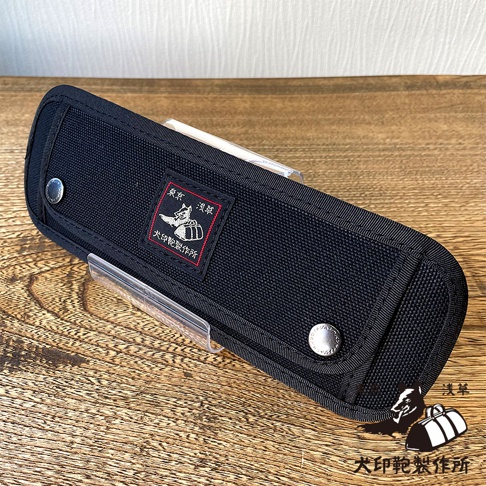 黑色純棉帆布製成的長形收納包，帶有兩個金屬按扣和一個紅色標籤，上面有白色字樣和圖案。