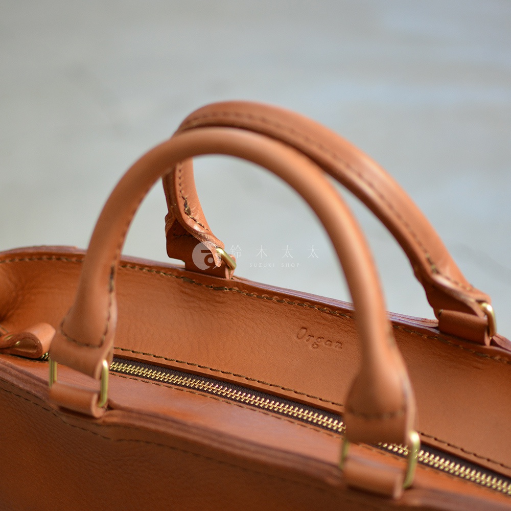 職人手作皮革Organ兩用商務托特包，呈現溫潤的棕褐色皮革質感，包款設有兩個圓弧形提把，搭配金色拉鍊與金屬配件，簡約而耐用。