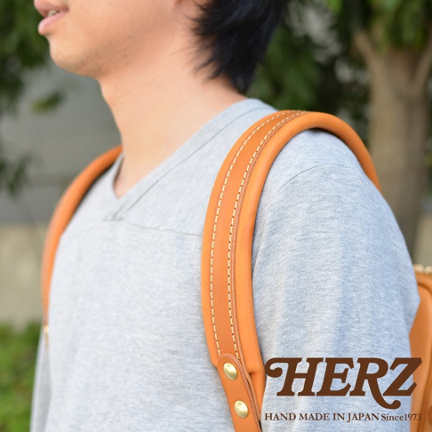 棕色皮革製成的雙肩背包，帶有縫線細節和黃銅扣件，一位男士穿著灰色上衣背著。