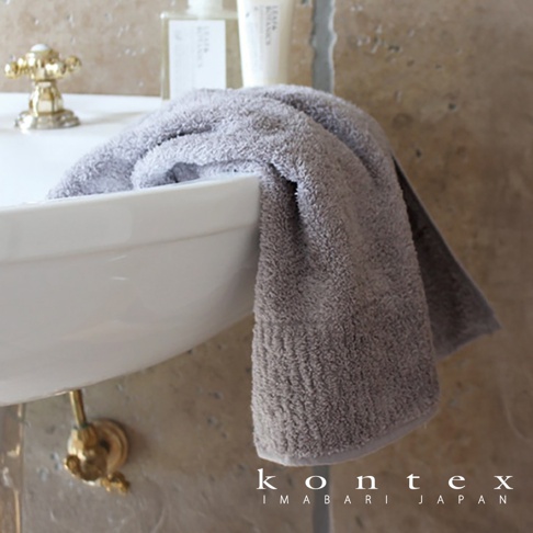 一條灰色極柔蓬鬆的今治毛巾，掛在白色洗手台上，背景是淺棕色紋理牆面和金色水龍頭。