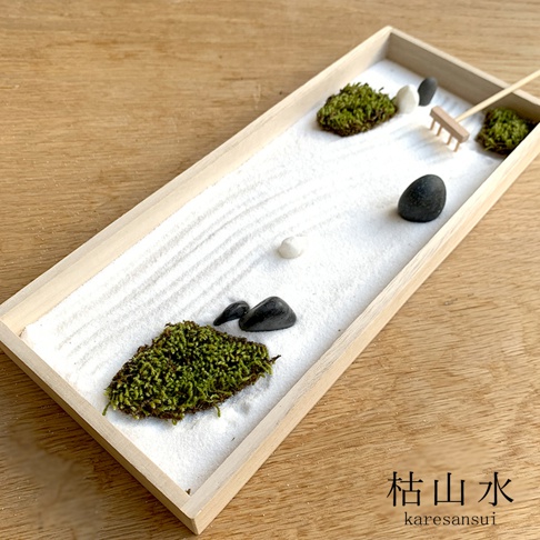 淺木色長方形盆中，舖有白色細沙，沙上耙出細緻紋路。盆內散落幾叢翠綠苔蘚、光滑的黑白卵石，以及一把迷你木耙，構成一幅日式枯山水景觀。