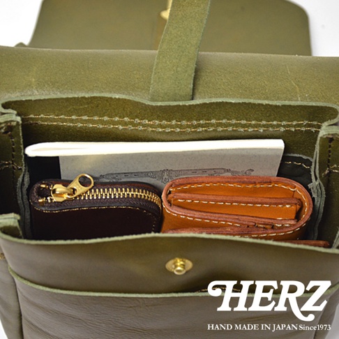 HERZ 品牌標誌與日本製造 since 1973 字樣，置於綠色皮革包的下方。包內可見一個棕色零錢包與一個棕色皮夾。