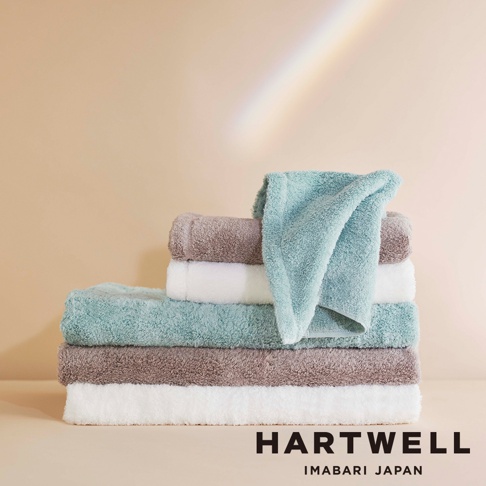 一疊棉質毛巾，包含淺藍綠色、白色和棕褐色，質地柔軟蓬鬆，頂部一條淺藍綠色毛巾斜放。