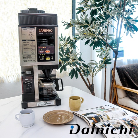 銀色與黑色金屬外殼的自動生豆烘焙咖啡機 MC-520A，配有玻璃咖啡壺、控制面板和咖啡豆儲存槽，置於白色桌面上，旁邊有兩杯咖啡，一個淺黃色馬克杯和一個白色咖啡杯，以及一個棕色盤子和一本打開的雜誌，背景是綠葉植物和窗簾。