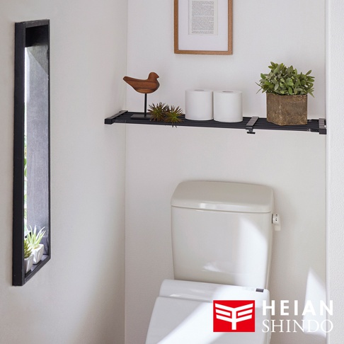 安裝在廁所上方牆壁的黑色兩段式強化伸縮桿，上面擺放著兩個捲筒衛生紙、一個木製小鳥裝飾、一叢綠色裝飾植物，以及一個盆栽植物。