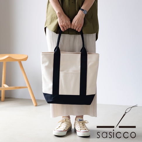 米白色三河木綿刺子織托特包，搭配黑色提把與底部，正面有一個黑色條紋裝飾的貼袋。