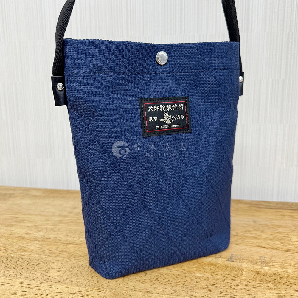 深藍色刺子織紋理平板收納側背包，正面有黑色標籤，印有金色日文及英文品牌資訊。包包配有黑色織帶和金屬按扣。