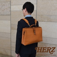 一位穿著西裝的男性背著一個棕色皮革的軟式杜勒斯包。包包為手提式，並配有可拆卸的肩帶，可轉換為背包模式。包包正面有一個扣環和綁帶設計，材質呈現自然皮革紋理。