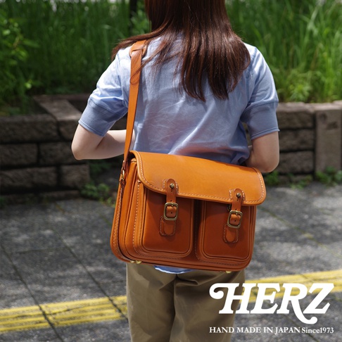 日本HERZ手工皮革雙扣郵差包，揹在身穿淺藍色上衣的女性肩上，採用焦糖色皮革製作，正面有兩個口袋和兩個金屬扣環。