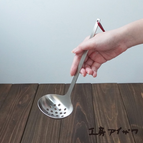 一只手正握著一把不鏽鋼萬用瀝水湯勺，勺面上佈滿圓形濾孔，勺柄末端有一個紅色掛繩。