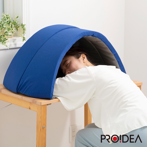 藍色防噪遮光舒眠罩，外層為柔軟布料，內部為灰色材質，呈現半圓形包覆設計，放置於木製桌面上，一位女性使用者將頭部置於其中休息。