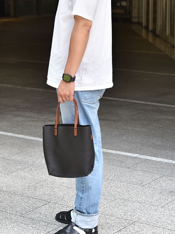 黑色軟皮直式托特包，搭配咖啡色皮革提把，由人手持於右側。