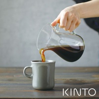 玻璃咖啡分享壺傾倒著深褐色咖啡液，注入一個灰色陶瓷馬克杯中。分享壺具有透明玻璃材質和彎曲的把手。