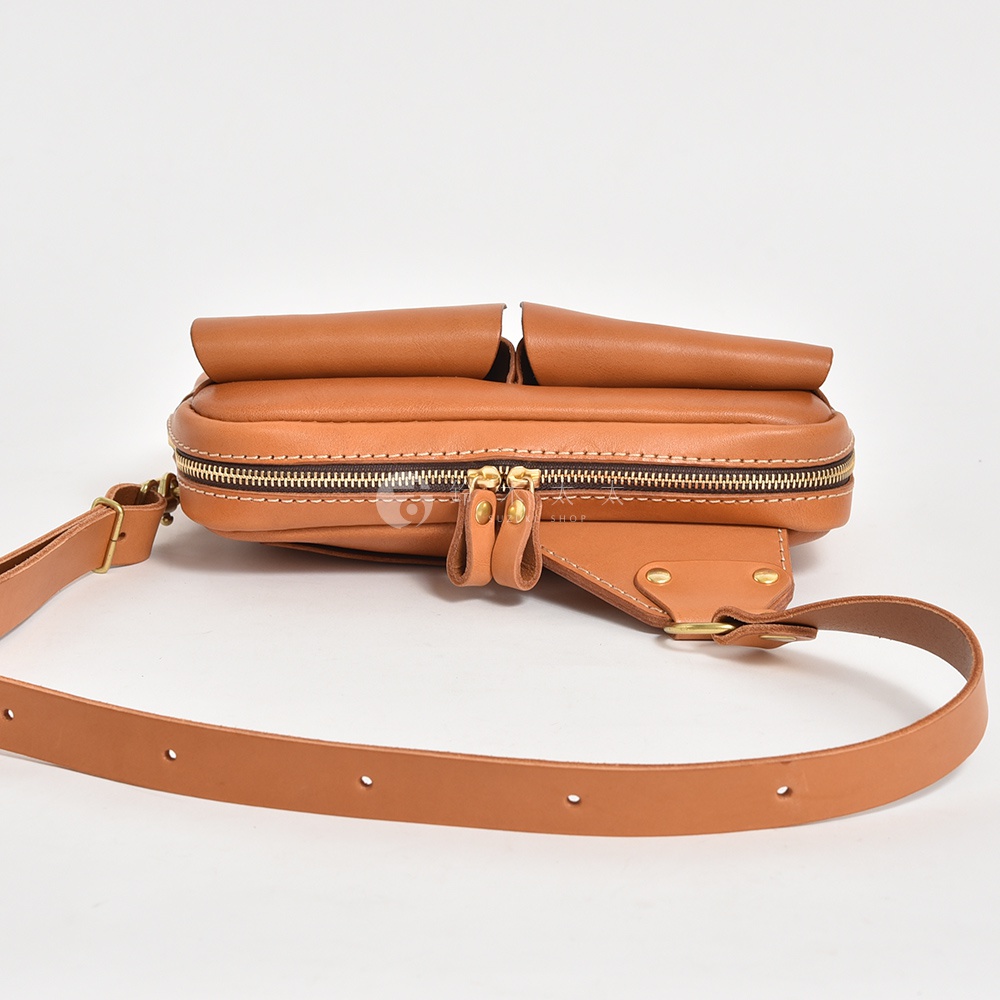 棕色皮革斜背包，有雙前袋設計，使用金色拉鍊和金屬配件，搭配可調節的皮革背帶。