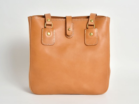 淺棕色皮革製的職人手作托特包，配有兩條短提把、一個中央磁扣封口，以及兩側的裝飾性壓扣設計。