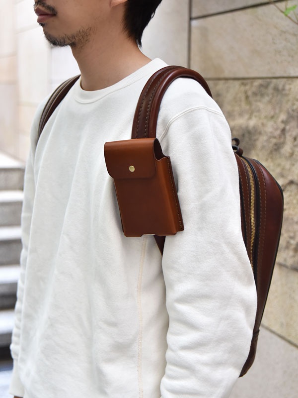 棕色皮革製成的三用手機收納包，附有金屬釦，固定於白色上衣肩帶上，旁邊可見同為棕色皮革的背包。