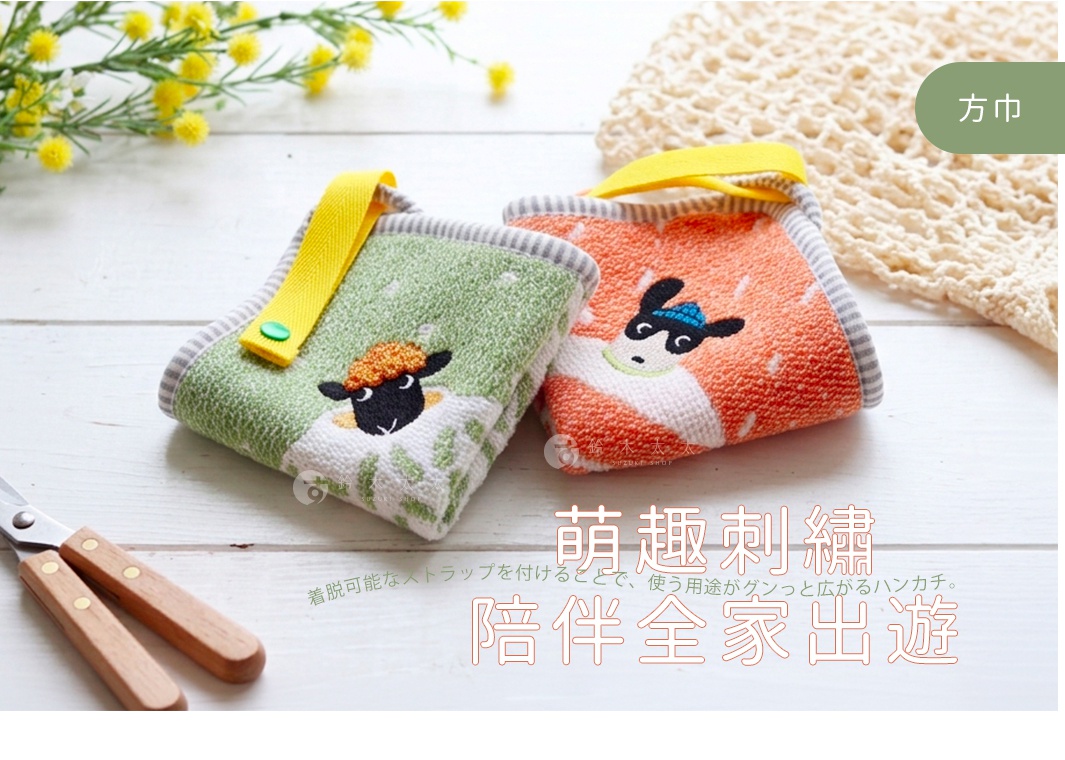 兩條帶有刺繡小羊圖案的今治純棉寶寶方巾並排放置於白色木紋桌面上，左側為綠色羊隻，右側為橙色羊隻，兩者上方皆附有黃色可拆卸式掛帶，背景裝飾有黃色小花與編織網袋。