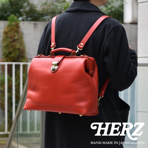 一位佩戴著日本HERZ品牌職人手作紅色皮革杜勒斯公事包的穿搭展示，包款呈現硬挺的方形結構，具備手提與後背雙用功能，金屬鎖扣與底部腳釘細節顯眼，右下角印有HERZ品牌名稱與標語。