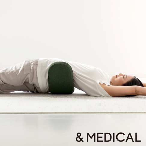 一位女性平躺在地上，將一個綠色的U型骨盤枕置於腰部下方。枕頭呈現半圓弧形，表面為織物材質。