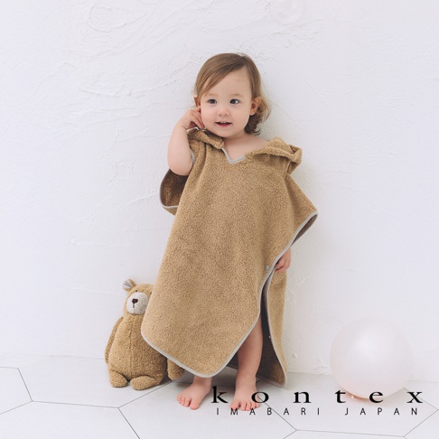 一名嬰兒穿著棕色連帽浴巾，浴巾帶有可愛的熊耳朵設計。浴巾材質柔軟，邊緣為灰色滾邊。一旁擺放著一隻泰迪熊玩偶和一個白色氣球。