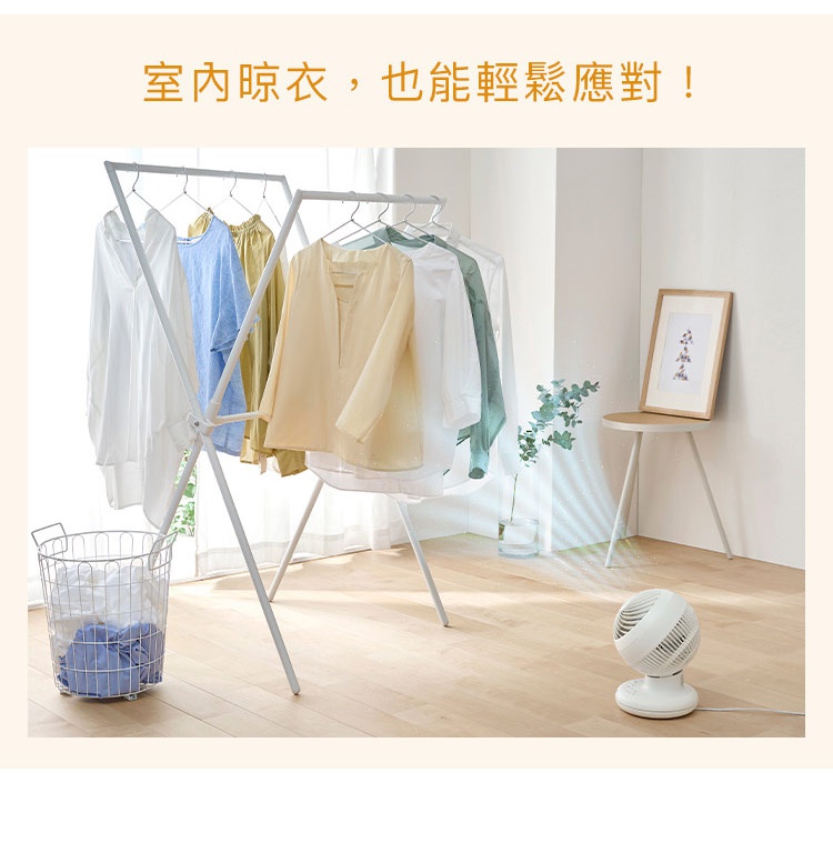 在明亮的室內空間中，架設著一座白色的折疊式晾衣架，上面掛滿了整齊的各色衣物。右下角放置著一台白色的圓形空氣循環扇，吹拂出模擬的氣流軌跡，指向晾衣架的方向，背景還有簡單的木製圓桌與裝飾畫，呈現出居家生活場景，上方文字標示室內晾衣也能輕鬆應對。